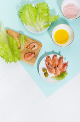 Cooking of spring salad with shrimps, green olive, greens, olive oil, ingredients on pastel minty background with shadow in hard light, top view, vertical.