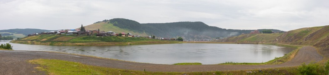 Small cozy village stands on shore of lake or river, hills and fog in background. Panoramic view. Early cool morning, overcast gray sky