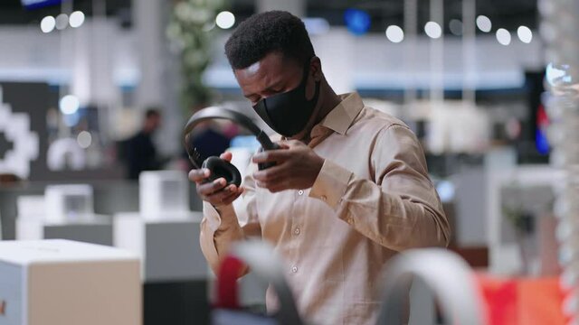 young black guy is viewing new model of wireless headphones in trading hall of audio equipment shop, medium portrait of male shopper