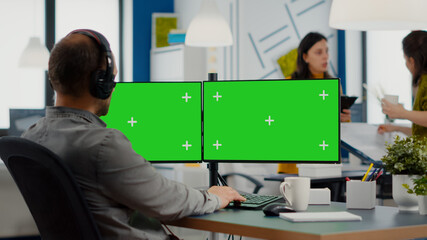 Employee with headphones using dual monitror setup with green screen, chroma key mock up isolated...