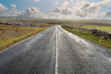 Empty narrow road in beautiful scenery. Burren Area, west of Ireland. Traditional stone fence and cloudy blue sky in the background. Wild Atlantic Way tourist route. Nobody. Day time