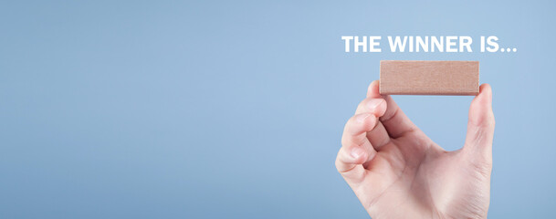 Male hand holding wooden block with a The Winner Is... text.
