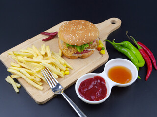 French fries, hamburgers on a wooden cutting board with a handle, near sweet sauce and green peppers, black background top view