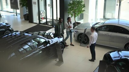 Young couple enters car salon and shakes hands with sales person, stands in light interior spbd. - Powered by Adobe