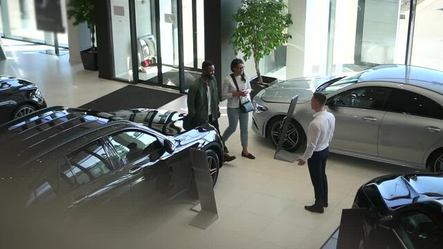 Young couple enters car salon and shakes hands with sales person, stands in light interior spbd.