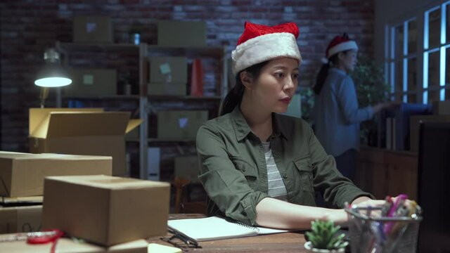 two asian korean women employee in internet shop working at dark office on Christmas eve. female e commerce worker busy for xmas gift shipping parcel delivery in studio. lady coworkers in santa hats