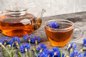 Cup of herbal tea, transparent teapot and blue cornflowers flowers on wood background. 
