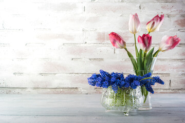 Flowers of tulips and grape hyacinths in a jar and crystal glass on a rustical brick wall with copy space
