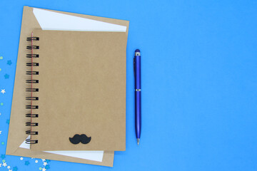 Layout of a greeting card for Father's Day.Blue envelope from a needlework notebook with a blue metal pen on a blue background. Fake black mustache in retro style.Top view.Flat styling style.Copyspace