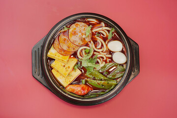 Spicy udon noodles in a earthenware pot on red background, eggs, sausages, greens, nutritious and delicious