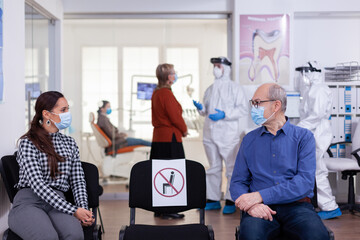 Senior man with face mask discussing with woman patient in stomatology clinic in waiting room, keeping social distancing during global pandemic with coronavirus. Doctor standing wearing ppe suit.