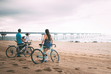 Friends seen from behind with their bikes on the beach at dawn looking out to sea. Young people...
