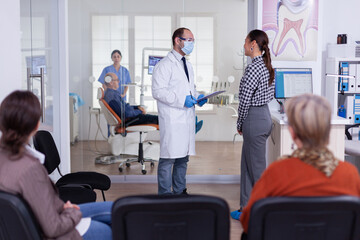 Dentist doctor interrogating woman and taking notes on clipboard standing in waiting area. Young patient explaining dental problem to stomatologist speaking in crowded reception of clinic. - obrazy, fototapety, plakaty