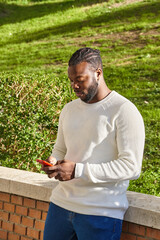 Fototapeta premium Handsome african american guy chatting via smartphone standing, cheerful dark-skinned man enjoying free time blogging in networks via smartphone.