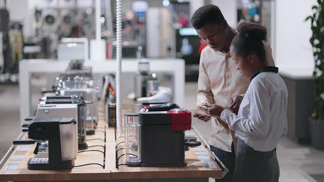 black man and woman are choosing automatic coffee maker in home appliances shop, reading manual of modern model