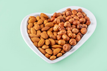 Assortment of various types of nuts - hazelnuts,  almonds in heart shaped plate on green background. Healthy vegetarian snacks. Protein-containing food. Close up.