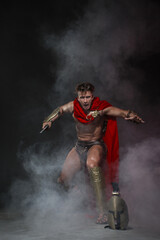 Young athletic sexy man in a costume of a Roman warrior in a red cloak in move on a black...