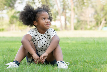 Happy little African child girl smiling and sitting on green grass with afro twin tails hair at park on summer.