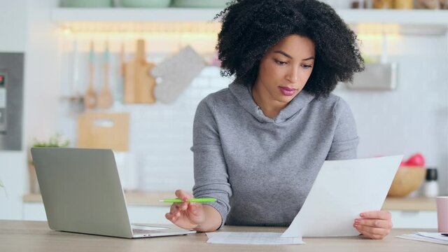 Video of afro business woman working with computer while consulting some invoices and documents in the kitchen at home.
