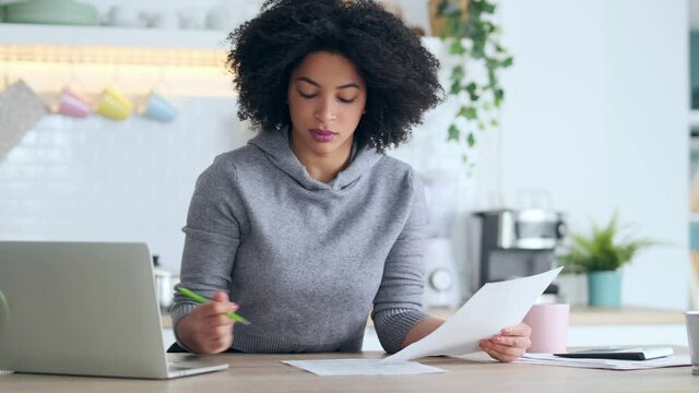 Video of afro business woman working with computer while consulting some invoices and documents in the kitchen at home.