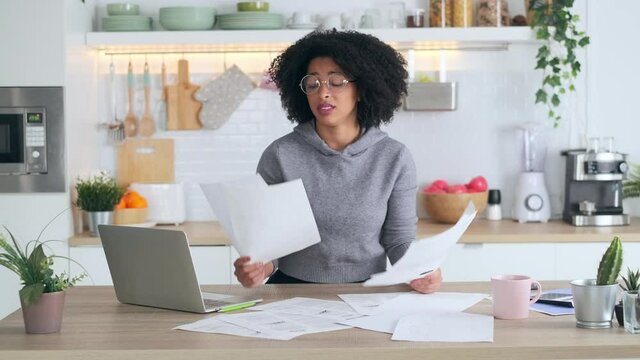 Video of bored afro business woman working with computer while consulting some invoices and documents in the kitchen at home.
