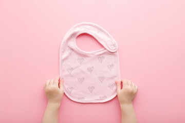 Baby girl hands holding textile bib with heart shapes for feeding on light pink table background. Pastel color. Closeup. Point of view shot. Top down view.