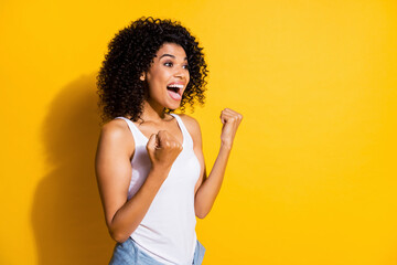 Photo of astonished young dark skin lady wear white singlet rising fists looking empty space isolated yellow color background