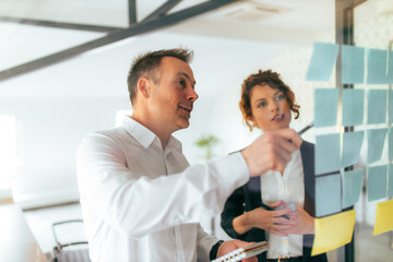 Discussing ideas. Two business people talking while looking at post it notes on a glass wall.