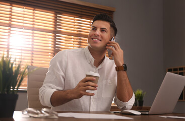 Freelancer with cup of coffee talking on phone while working at table indoors