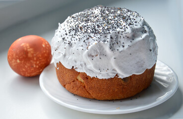 Easter cake is watered with white glaze and sprinkled with poppy seeds on top. One brown chicken egg dyed with onion skins. Close-up photo on a light blue background.