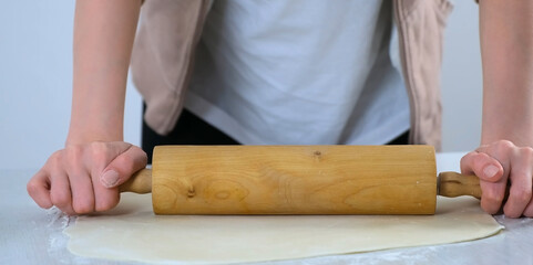 Woman is rolling dough for dumpings using rolling pin on table in kitchen, hands closeup. Process of cooking dumplings at home. Minced meat, dough and rolling pin on the table.