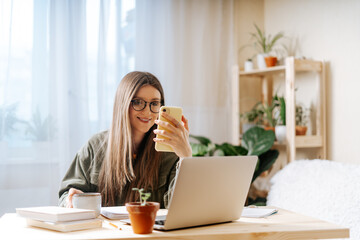 Freelance woman in glasses with mobile phone typing at laptop and working from home office with plants. Happy girl on workplace at the desk taking selfie. Distance learning online education and work.