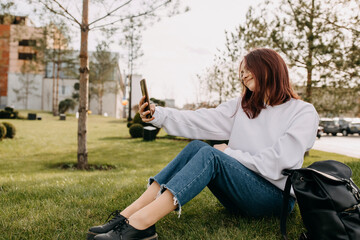 Young happy female student sitting on grass in park, with a backpack, smiling and taking a selfie...