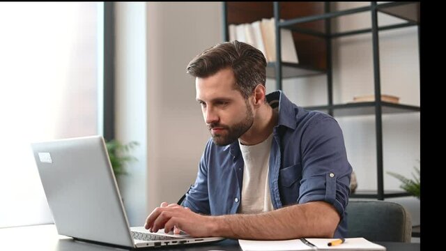 Handsome bearded male entrepreneur in casual wear using laptop for remote work from home, concentrated freelancer guy typing, looks through sheets of contract, sending emails, cheks report