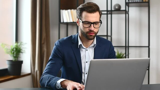 Focused concentrated determined young bearded office worker in business formal wear, sitting at the desk, typing on laptop, businessman professional is working on project, writing email to client