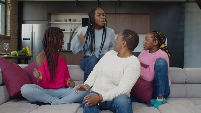 Emotionally stressed adorable african american teenage sisters loudly arguing on sofa, expressing complaints, mutual offends and frustration while caring pretty mother trying to calm down conflict.