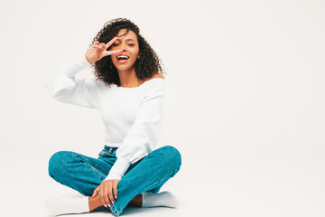 Fototapeta premium Beautiful black woman with afro curls hairstyle.Smiling model in sweater and trendy jeans clothes. Sexy carefree female posing on white background in studio. Tanned and cheerful.Shows peace sign