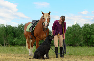 Caucasian mature woman, 36 years old, her horse and her dog are standing on a field.