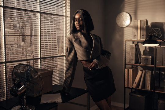 beautiful afro-american girl in a gray short jacket and black short skirt stands at a table in the office