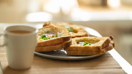 Morning breakfast. Fragrant baked sandwiches with egg, ham, cheese and pickles lie on a plate on the table, next to a mug of green tea in the kitchen on the table with morning sunlight.