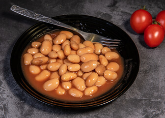White falo in tomato sauce. Boiled beans, stewed with tomato sauce in a black plate on a dark surface and cherry tomatoes. Close-up