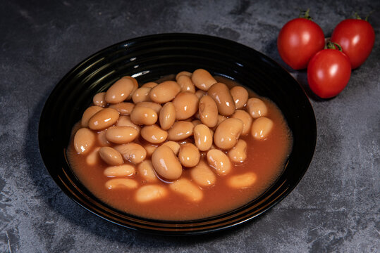 White falo in tomato sauce. Boiled beans, stewed with tomato sauce in a black plate on a dark surface and cherry tomatoes. Close-up