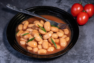 White falo in tomato sauce. Boiled beans, stewed with tomato sauce in a black plate on a dark surface and cherry tomatoes. Close-up