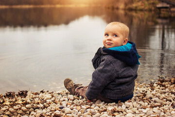 Little caucasian kid is sitting on the beach of the lake in spring or autumn nature.