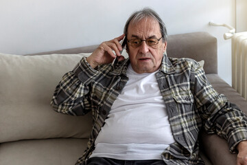 Obraz premium Elderly man in casual clothes seated on sofa in living room holding mobile phone, having distant communication. Retired hispanic man talking on cell sitting at home. Senior man receiving unwanted call