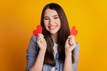 Close-up of young romantic affectionate woman hold two little hearts on yellow background