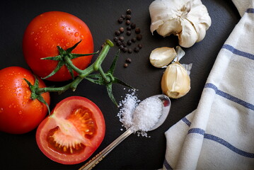 Fresh tomatoes, garlic, salt, pepper, kitchen cloth on a black table, top view