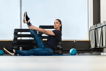 Fototapeta na wymiar Women and sport. A beautiful girl in sports clothes does stretching in the gym, pulls the leg up and listening to music in wireless headphones. Healthy lifestyle concept