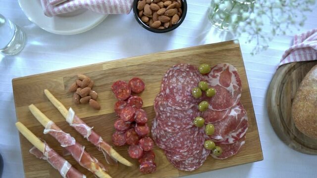 Looping top down footage pans across sharing party meat platter and snack bowls including olives, nuts and cheese. Meal sharing, charcuterie board food concept in flat lay style with natural lighting