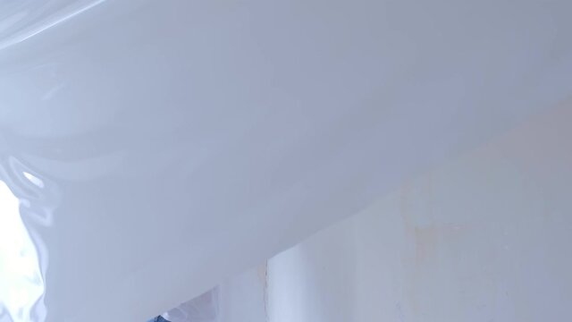 Man builder dismanting of stretch suspended ceiling cuts it with knife. He is looking at camera, smiling peeking out from under fabric. Building construction work. Process of removing and renovation.
